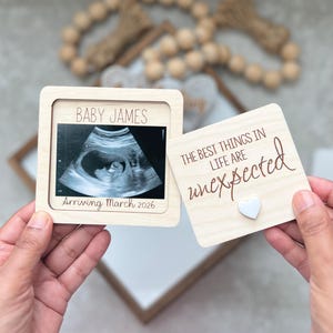 May include: A wooden pregnancy announcement with the text "BABY JAMES" and "Arriving March 2026" around an ultrasound image. A separate wooden square reads "THE BEST THINGS IN LIFE ARE unexpected" with a small white heart.