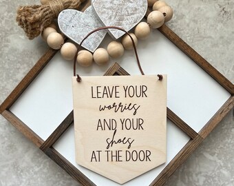May include: Wooden sign with the text "Leave your worries and your shoes at the door". The sign is hanging from a brown leather cord and is decorated with a wooden bead garland and two white wooden hearts.