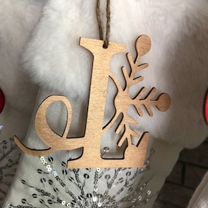 May include: Wooden Christmas ornament in the shape of the letter "L" with a snowflake design. The ornament is made of light brown wood and hangs from a natural twine string. The background features a white stocking with sequins.