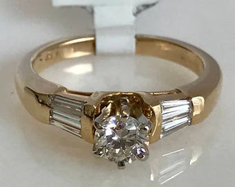 May include: A gold engagement ring with a round center diamond and two baguette diamonds on either side. The ring is set in a simple band.