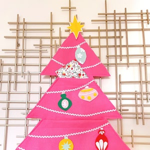 May include: A pink felt Christmas tree with a white scalloped edge and a variety of felt ornaments, including a star, a bell, and a candy cane. The tree is standing on a gold metal grid.