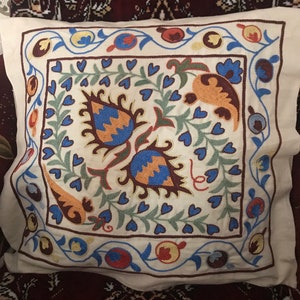 Op de afbeelding: Crèmekleurig vierkant kussen met ingewikkelde geborduurde bloemenpatronen. De borduursels tonen blauwe, oranje en groene bloemen en bladeren, omlijst door een rand met blauwe en rode accenten. Een decoratief woondecoratie-item.