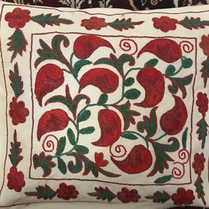 Op de afbeelding: Een wit kussen met rood en groen geborduurde bloemenmotief. Het kussen heeft een vierkante vorm en is versierd met een herhalend patroon van gestileerde bloemen en bladeren.