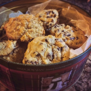 May include: A tin of assorted cookies, including chocolate chip and oatmeal raisin, sits inside a red and green plaid tin. The cookies are light brown and golden, with visible chocolate chips. The tin is lined with parchment paper.