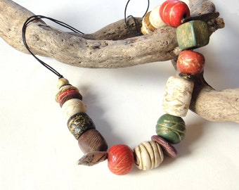 Puede incluir: Un collar artesanal con cuentas coloridas en rojo, verde, crema y marrón. Las cuentas están ensartadas en un cordón negro y descansan sobre un trozo de madera flotante. El collar tiene un diseño artístico único.