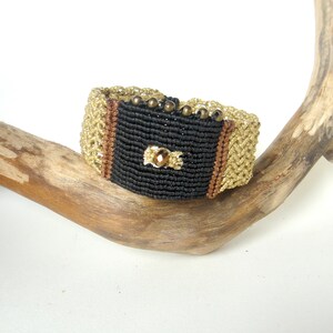 May include: A handmade macrame bracelet featuring a central black panel with a decorative gold-colored jewel. The bracelet has gold and brown accents, and is adorned with small, bronze-colored beads. The bracelet is displayed on a piece of driftwood.