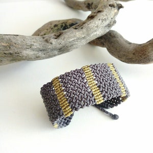 May include: A handmade macrame bracelet in gray and gold. The bracelet features a woven design with alternating stripes of gray and gold thread. The bracelet is adjustable and appears to be made of cotton or a similar material.
