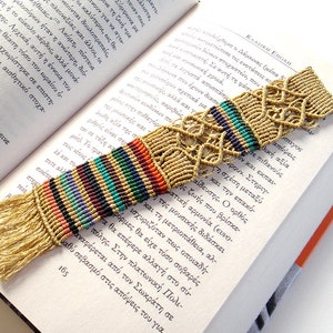 Pode incluir: Um marcador de livros de macramé feito à mão com uma base bege e uma seção listrada colorida. O marcador de livros tem uma extremidade com franjas e é colocado em um livro aberto. O marcador de livros tem cerca de 20 cm de comprimento.