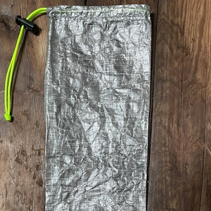 Könnte beinhalten: Eine silberne Kordeltasche mit einem leuchtend gelben Kordelzug. Die Tasche besteht aus einem leichten, zerknitterten Material.