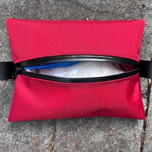 Op de afbeelding: Een rechthoekig rode etui met een zwarte ritssluiting en zwarte stoffen lussen aan elk uiteinde. De ritssluiting is gedeeltelijk open en onthult witte en blauwe inhoud. De etui is gemaakt van een textuurrijk materiaal.