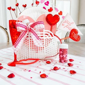 May include: A white wire basket filled with Valentine's Day themed gifts, including a red heart lollipop, a pink and white gingham bow, a red lip-shaped glass, and a small jar of red and pink heart sprinkles. The basket is sitting on a white and pink striped table with red heart confetti scattered around it.
