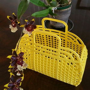 May include: A yellow plastic tote bag with a grid pattern. The bag has two handles and is sitting on a dark brown surface. A brown and yellow orchid is in the background.