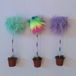 May include: Three decorative potted plants featuring tall, thin, speckled stems topped with round, fluffy faux fur pom-poms. The left plant has mint green fur, the center has multi-colored rainbow fur, and the right has light purple fur.