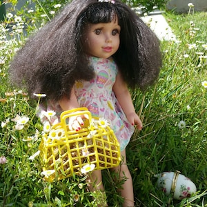 Puede incluir: Una muñeca con el pelo largo y oscuro que lleva un vestido rosa y blanco sostiene una cesta amarilla llena de flores blancas. La muñeca está de pie en un campo de hierba verde y flores blancas.