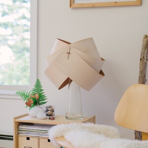 May include: A wooden lamp with a unique, folded shade sits on a wooden side table with drawers. The lamp is next to a vase with a fern and a glass jar with decorative stones. A white sheepskin rug sits on a wooden chair.