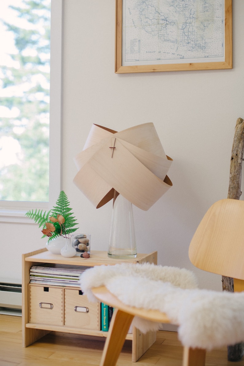 May include: A wooden lamp with a unique, folded shade sits on a wooden side table with drawers. The lamp is next to a vase with a fern and a glass jar with decorative stones. A white sheepskin rug sits on a wooden chair.