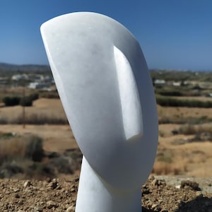 Op de afbeelding: Een witte marmeren sculptuur van een gestileerde kop met een gladde, gebogen vorm. De sculptuur heeft een eenvoudig, elegant ontwerp met een prominente neus en een afgeronde bovenkant. Het kunstwerk staat tegen een achtergrond van een blauwe lucht en een natuurlijk landschap.