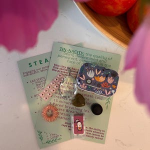 May include: A selection of self-care products, including a patterned tin, ear seeds, a finger massager, and a card with text. The items are displayed on a white surface, with a blurred background of pink flowers and apples.