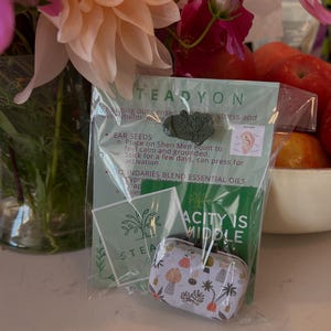 May include: A clear plastic package containing a small metal tin with a colorful botanical pattern, a green card with the text "ACITY IS MIDDLE", and a green stone with a tree design. The package is in front of a bouquet of flowers.