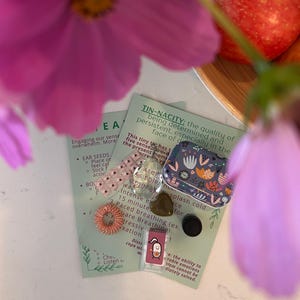 Puede incluir: Una colección de artículos de bienestar que incluye una pequeña lata con estampado floral, una hoja de semillas para los oídos rosa, un masajeador de dedos y una piedra en forma de corazón. La imagen también muestra texto sobre la participación de los sentidos y el manejo de la sobrecarga.