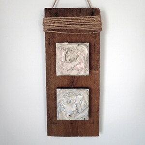 May include: Two square ceramic tiles with abstract swirling patterns in pink and white and blue and white are mounted on a rustic wooden plank. The plank is hung on a white wall with twine.