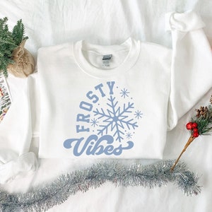 Puede incluir: Sudadera blanca con un diseño de copo de nieve azul y el texto "FROSTY VIBES" en una fuente retro.