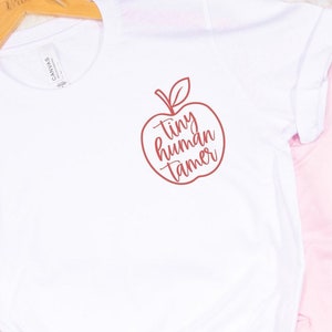 May include: White t-shirt with a red apple graphic and the text "tiny human tamer" inside the apple.