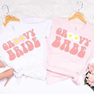 May include: Two white t-shirts with pink text and flower graphics. One shirt says "Groovy Bride" and the other says "Groovy Babe".