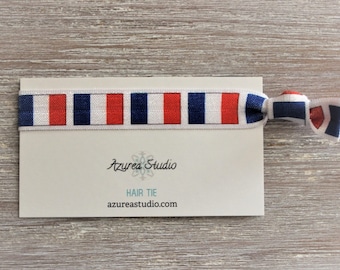 May include: A red, white and blue striped hair tie with a white background. The hair tie is tied in a bow and has the text "Azurea Studio" and "Hair Tie" on a white card.