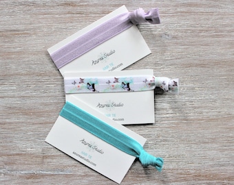 May include: Three hair ties on white cards with the text "Azurea Studio" and "Hair Tie" on each card. The hair ties are purple, white with a butterfly pattern, and turquoise.