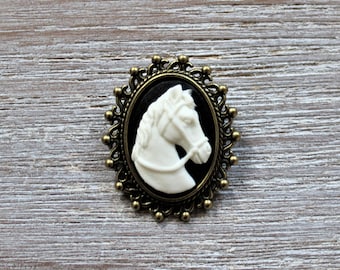 May include: A white cameo brooch featuring a horse's head in profile, set in a black oval with an ornate gold-toned frame.