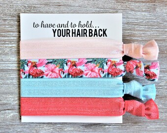 May include: Set of four hair ties with a flamingo and tropical leaf print, a solid pink hair tie, a solid light blue hair tie, and a solid coral hair tie. The hair ties are packaged on a white card with the text "to have and to hold... YOUR HAIR BACK".