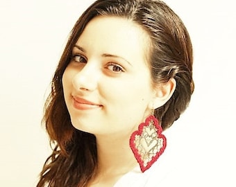 May include: A woman with long brown hair wearing a red and gold dangle earring. The earring is a delicate, intricate design with a heart shape.