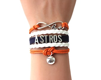 May include: A multi-strand bracelet with the word "ASTROS" in silver letters on a navy blue band. The bracelet includes orange and white braided cords, a baseball charm, and an infinity symbol with the word "love".