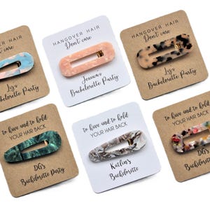 May include: Six brown cards with white text and a variety of patterned hair clips. The text on the cards reads "Hangover Hair Don't Care" and includes the names of the bachelorette party attendees. The cards also include the phrase "To have and to hold your hair back".