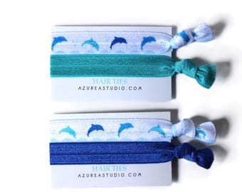 May include: Two sets of hair ties featuring a dolphin design. The top set has teal and white hair ties, and the bottom set has blue and white hair ties. The card reads "Hair Ties" and "azureastudio.com".