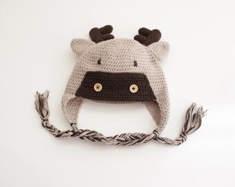 May include: A brown and beige crocheted hat with a reindeer design. The hat has two brown buttons for eyes and a long braided cord.