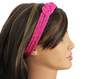 May include: A bright pink headband with a braided rope design. The hair accessory is worn by a person with brown hair. The braided rope detail adds texture and visual interest. The background is plain white.