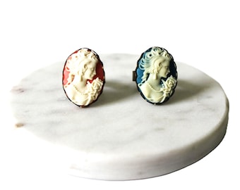 May include: Two cameo rings with a white cameo of a woman's profile. One ring has a red background and the other has a blue background.