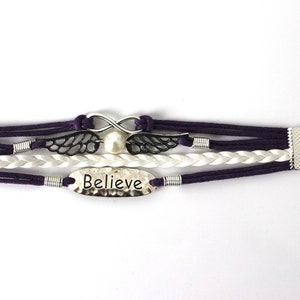 May include: A multi-strand bracelet with purple and white cords, a silver infinity symbol, a pearl, and a silver charm with the word "Believe" on it. The bracelet has a silver clasp and chain.