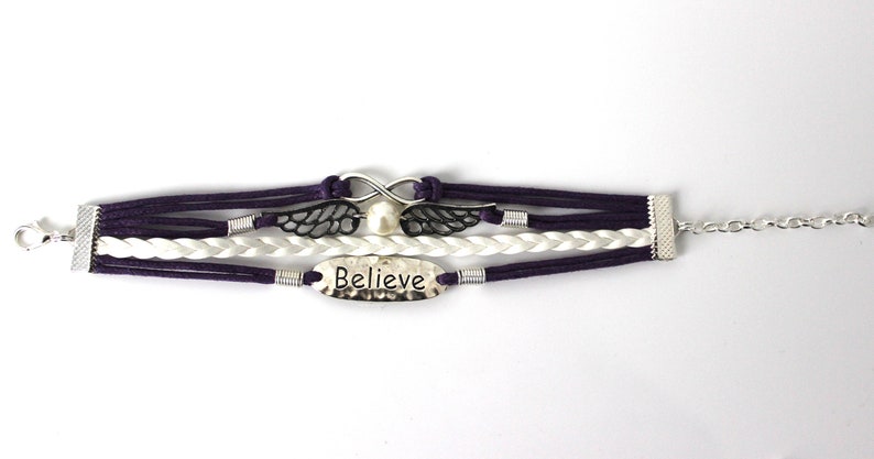 May include: A multi-strand bracelet with purple and white cords, a silver infinity symbol, a pearl, and a silver charm with the word "Believe" on it. The bracelet has a silver clasp and chain.