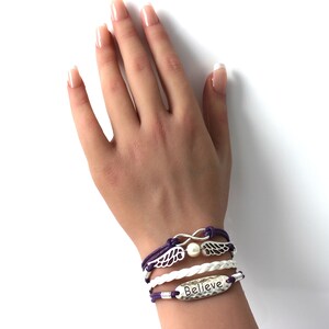 May include: A layered bracelet set with a white braided cord, purple cord, and a silver charm with wings and an infinity symbol. The bracelet also features a silver charm with the word "Believe" on it.