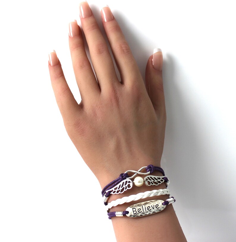 May include: A layered bracelet set with a white braided cord, purple cord, and a silver charm with wings and an infinity symbol. The bracelet also features a silver charm with the word "Believe" on it.