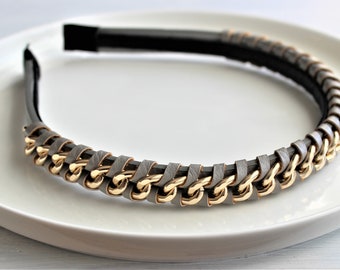 May include: A gray headband with a gold chain design. The headband features a black base and is decorated with a series of gold-colored chain links, interwoven with gray leather-like material. The accessory is displayed on a white plate.