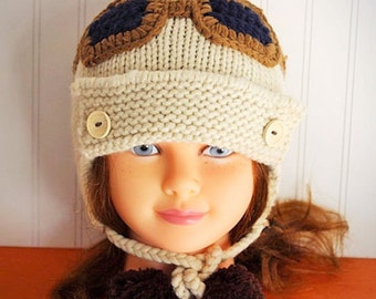 May include: A white knitted hat with brown and blue aviator goggles, two brown pom-poms, and two buttons. The hat has ear flaps that tie under the chin.