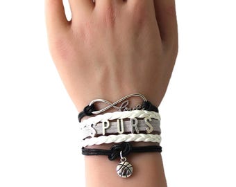 May include: A white and grey braided bracelet with a silver infinity charm that says "Love" and a silver basketball charm. The bracelet also has a silver charm with the word "SPURS" on it.