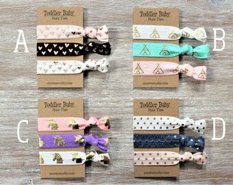 May include: Four sets of three elastic hair ties for toddlers. Each set features a different pattern: pink with gold hearts, teal with gold tipis, pink with gold unicorns, and navy blue with gold polka dots.