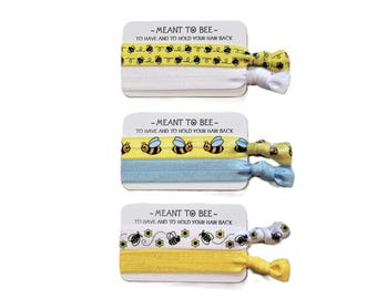 May include: Three sets of hair ties with bee-themed designs. Each set includes a card with the text "-MEANT TO BEE- TO HAVE AND TO HOLD YOUR HAIR BACK." The hair ties feature yellow, white, and blue bands with bee and flower patterns.