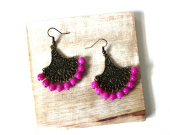 May include: A pair of ornate, antique bronze-colored earrings with a fan-shaped design. Each earring features a row of vibrant pink beads along the bottom. The earrings are displayed on a light-colored wooden surface.