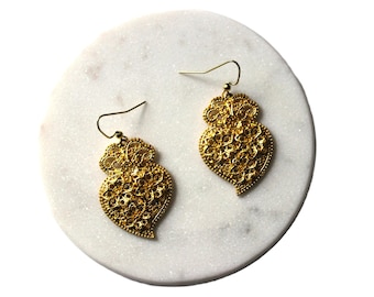 May include: A pair of gold filigree heart-shaped earrings with a delicate design. The earrings are hanging from a white marble surface.